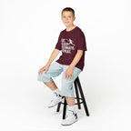 Young boy sitting on a stool wearing a purple t-shirt with a colorful graphic and text, light blue shorts, and white sneakers.