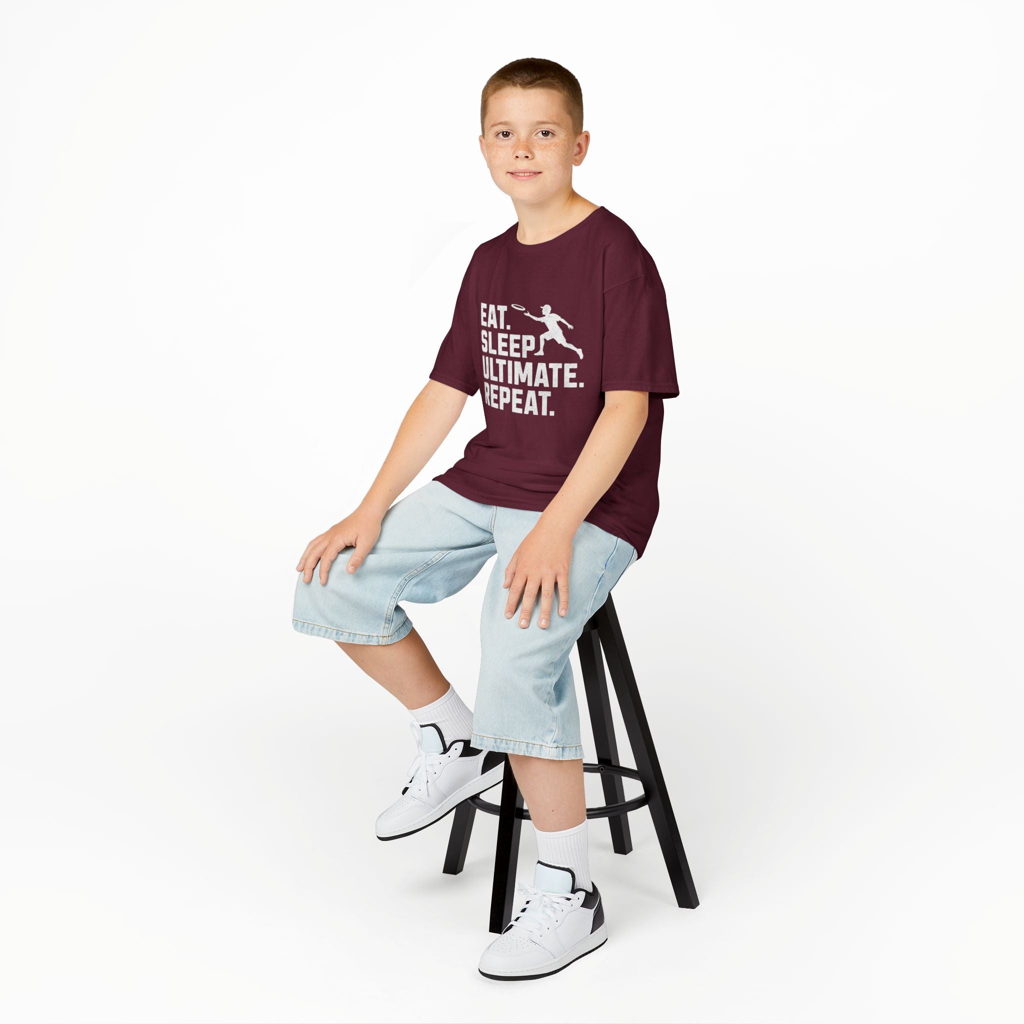 Young boy sitting on a stool wearing a purple t-shirt with a colorful graphic and text, light blue shorts, and white sneakers.