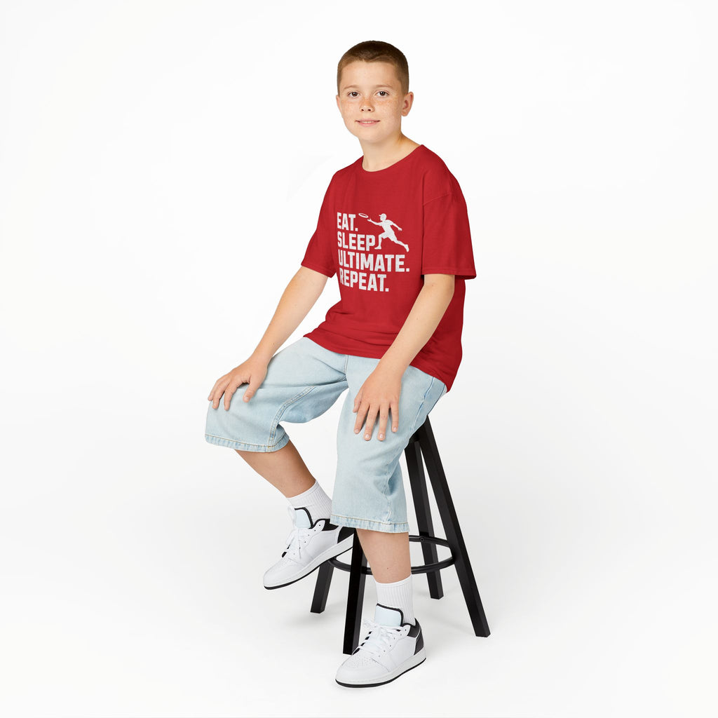 Child sitting on a stool wearing a red t-shirt with a graphic design and light blue shorts on a white background.