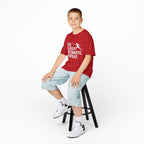 Child sitting on a stool wearing a red t-shirt with a graphic design and light blue shorts on a white background.