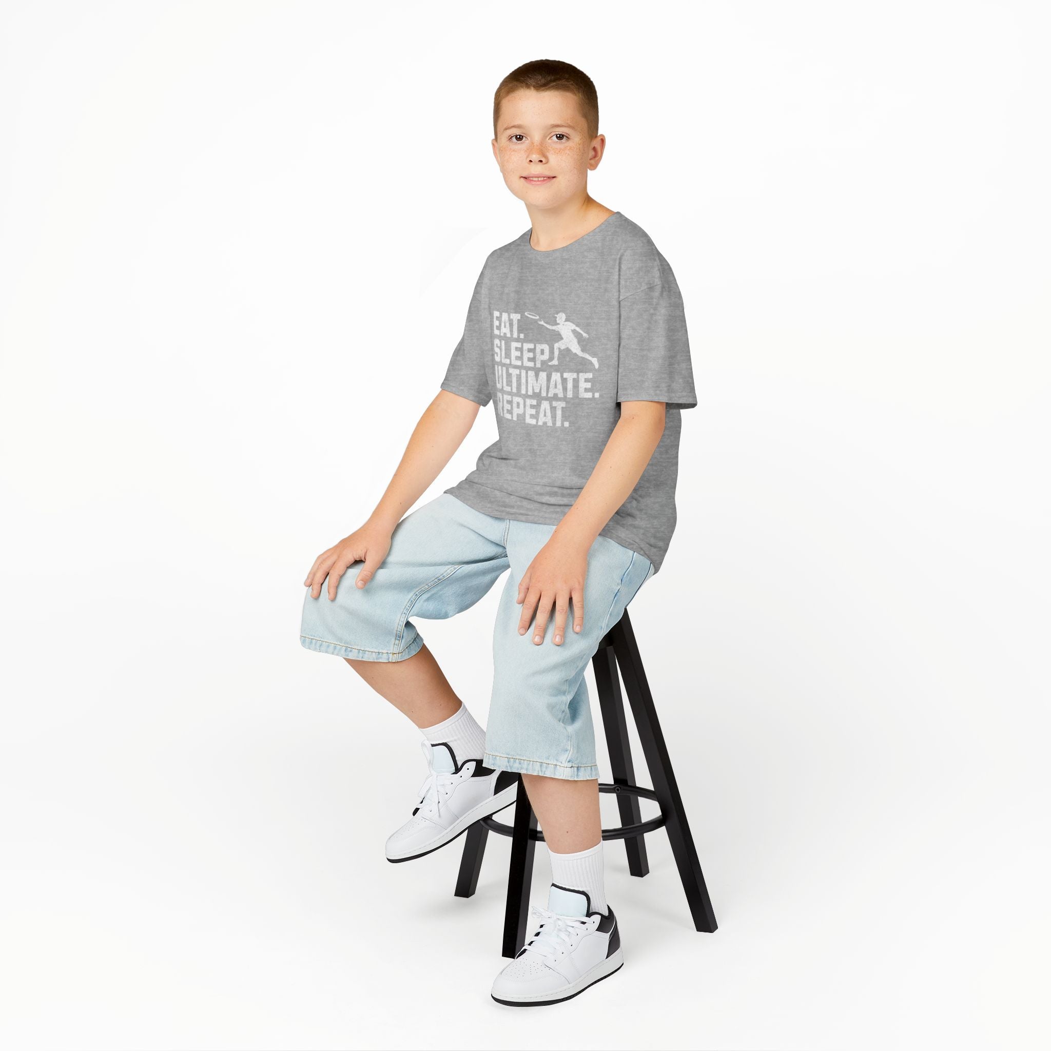 Child sitting on a stool wearing a gray t-shirt with text and light blue shorts on a white background