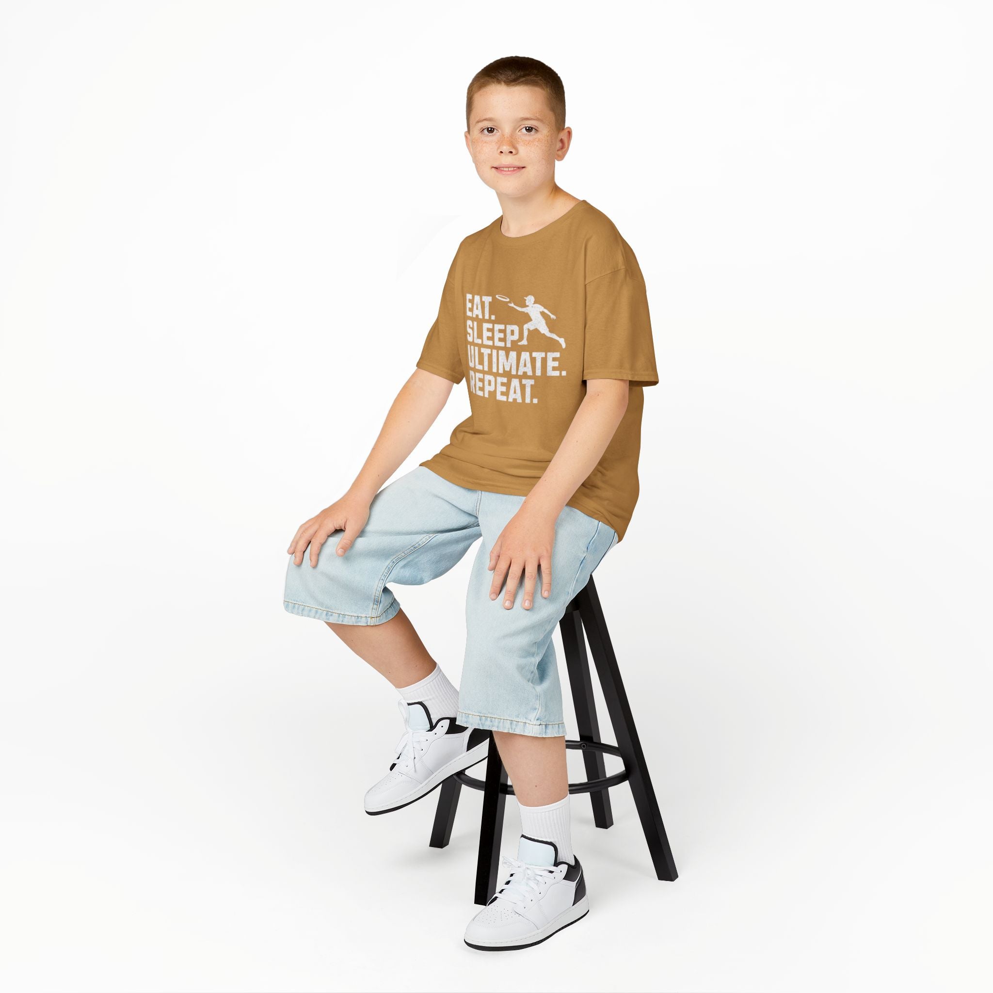 Child sitting on a stool wearing a brown t-shirt with text and light blue shorts on a white background