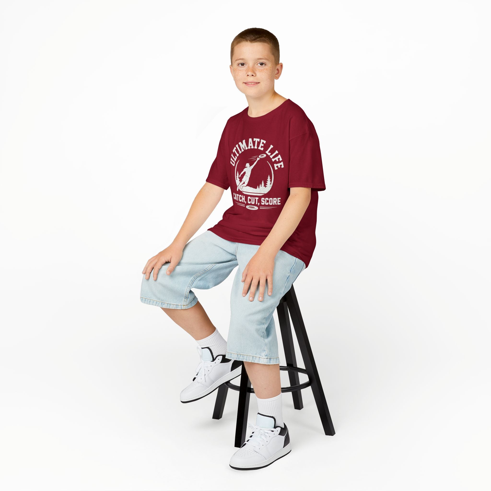 Young boy sitting on a stool wearing a red t-shirt with text and graphics, light blue jeans, and white sneakers.