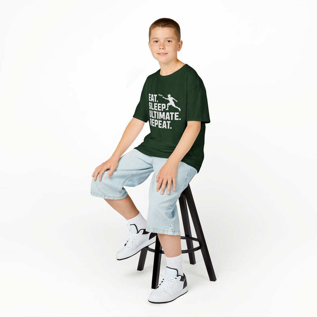Young boy sitting on a stool wearing a green t-shirt with text and light blue shorts on a white background