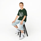 Young boy sitting on a stool wearing a green t-shirt with text and light blue shorts on a white background