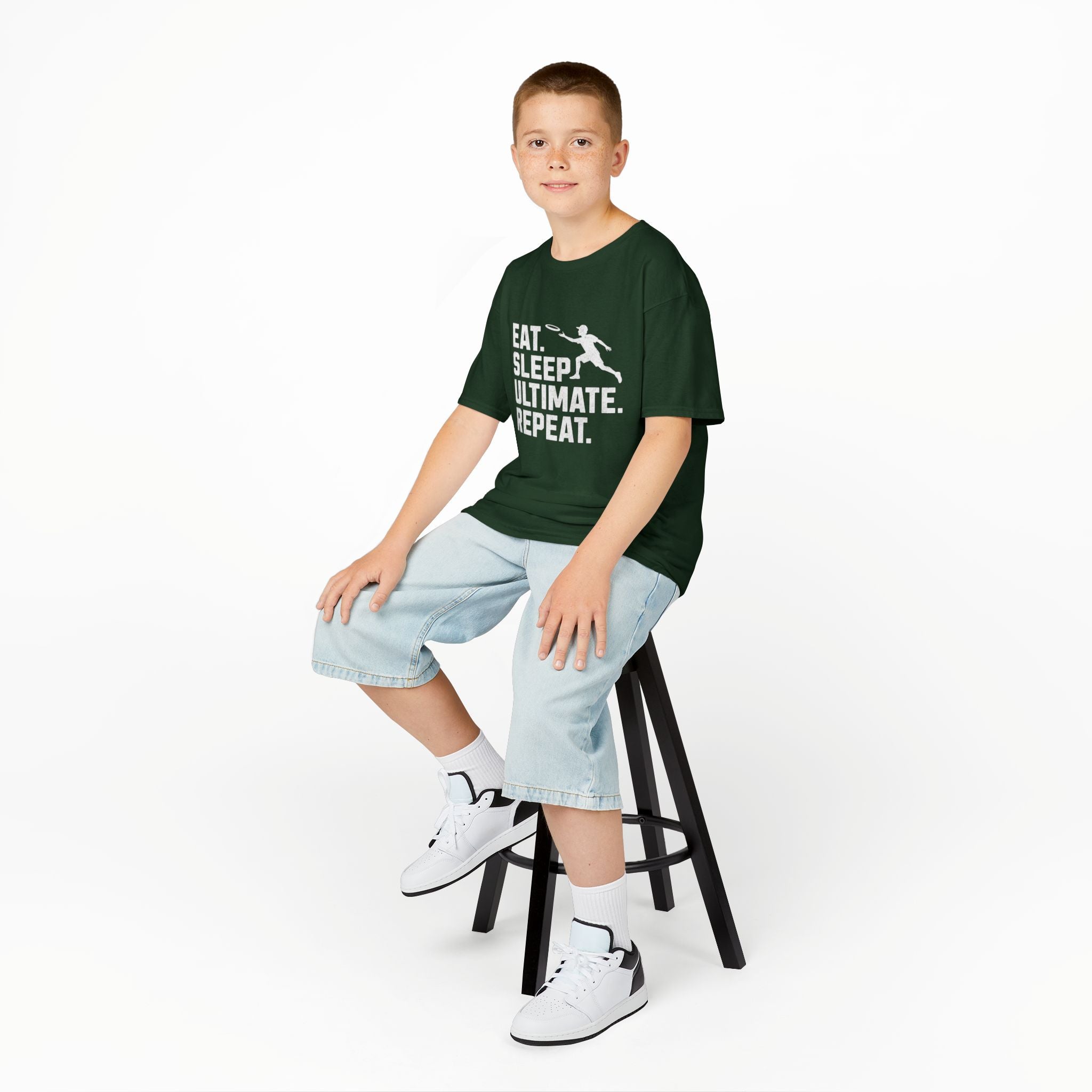 Young boy sitting on a stool wearing a green t-shirt with text and light blue shorts on a white background