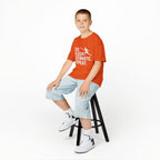 Child sitting on a stool wearing an orange t-shirt with text and graphics, light blue shorts, and white sneakers.