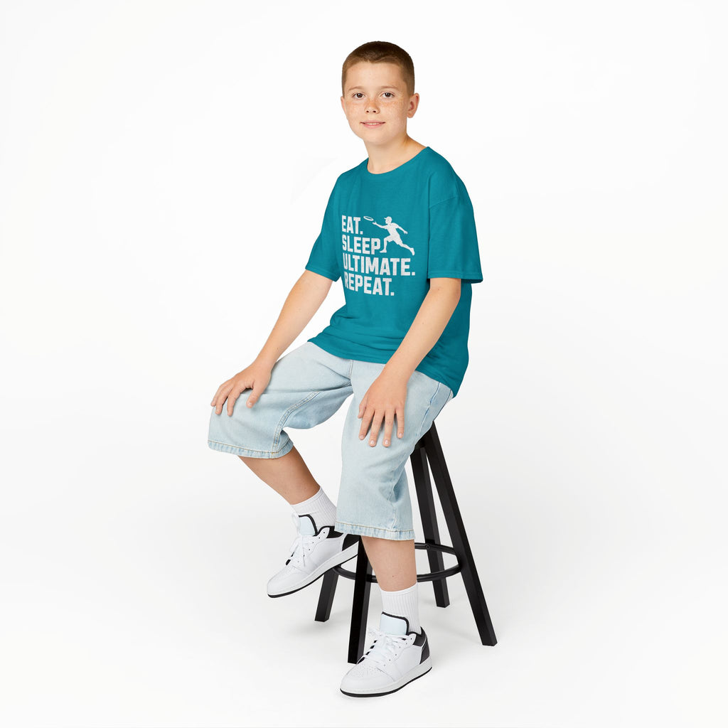 Child sitting on a stool wearing a teal t-shirt with text and light blue shorts on a white background
